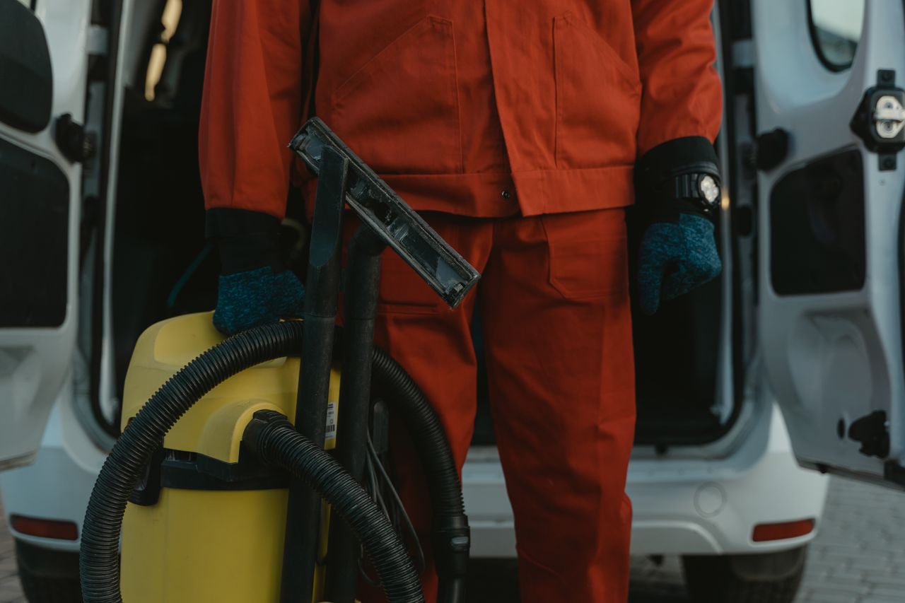 services-01 Un agent d’entretien professionnel en uniforme rouge tient un aspirateur près d’un véhicule utilitaire, prêt à intervenir.